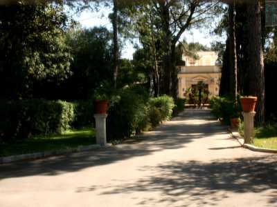 Foto 6b. Le sbarre del cancello sono coperte da una fittissima rete da cui riusciamo comunque a intravedere un breve sentiero alberato che conduce a una residenza di un delicato color ocra, al cui centro spicca un portone timpanato 